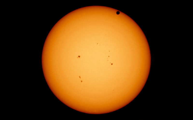 Fotografie der Sonne. Im Zentrum am hellsten dunkelt die gelbe Kreisscheibe zum Rand hin ab. Am oberen Rand klein der kreisrunde schwarze Schatten vom Merkur.