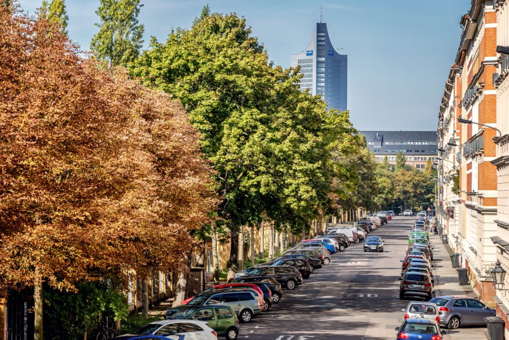 Blick entlang einer Straße. Die Hälfte des Bildes ist eingenommen von Straßenbäumen am linken Straßenrand. Fluchtpunkt in rechter Bildhälfte auf mittlerer Höhe. Häuserzeile am rechten Straßenrand. Straße voll geparkt mit Autos.