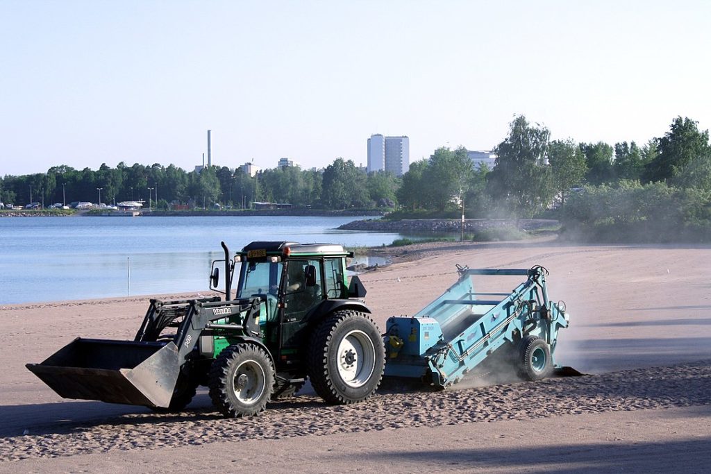 Ein Traktor zieht einen Sandreiniger über einen Sandstrand. Links eine Bucht. Im Hintergrund Bäume. Dahinter ragen zwei Hochhäuser und ein Schornstein in den Himmel.