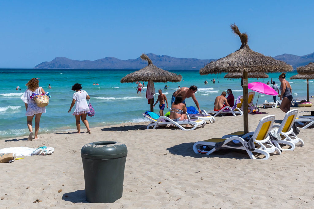 Grauer Mülleimer aus Plastik am Sandstrand. Strohsonnenschirme, Liegen, Personen. Im Hintergrund das Meer un am Horizont Berge.