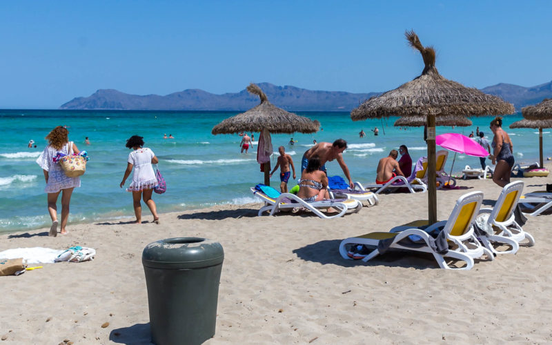 Grauer Mülleimer aus Plastik am Sandstrand. Strohsonnenschirme, Liegen, Personen. Im Hintergrund das Meer un am Horizont Berge.