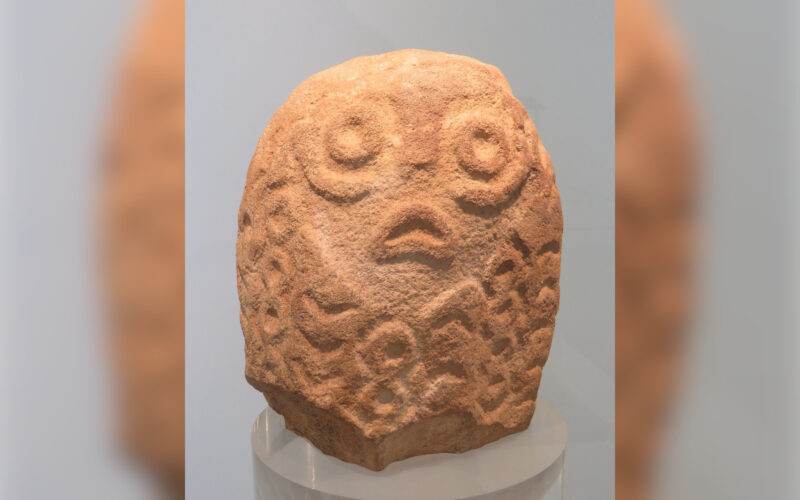 Hellbraune, ovale Steinfigur. Glattes Gesicht mit kreisrunden Augen, Nase und herabgezogenen Mundwinkeln. Rest der Figur mit Kreisen und Winkeln überzogen.