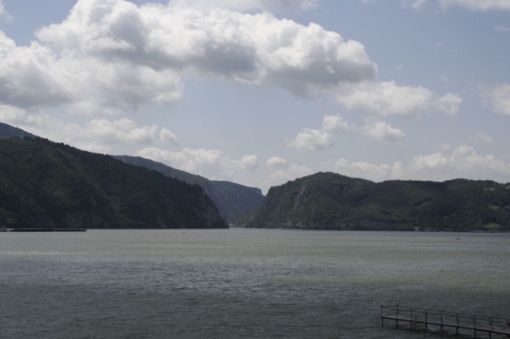 Blick entlang eines Flusses, der sich über die gesamte Breite des Bildes erstreckt. In der Mitte der Durchbruch zwischen Bergen. Im Hintergrund blauer Himmel mit vereinzelten Quellwolken.